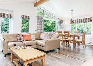 A modern living room with a sofa, dining table, and chairs, large windows with curtains, and a wooden floor. - Netherfield Watersedge Lodge (Newton Saint Loe, Nr Bath)