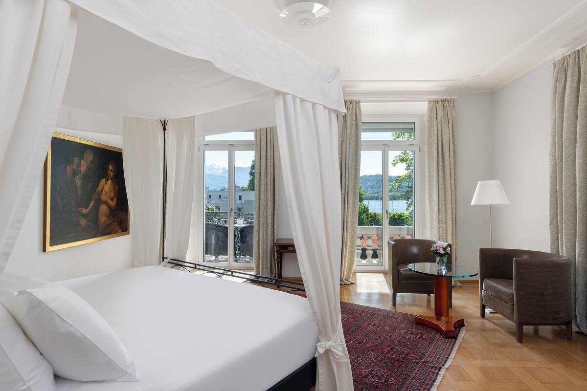 Ein Schlafzimmer mit einem Bett, einem Gemälde, einem Tisch mit einer Vase und Blick ins Freie.