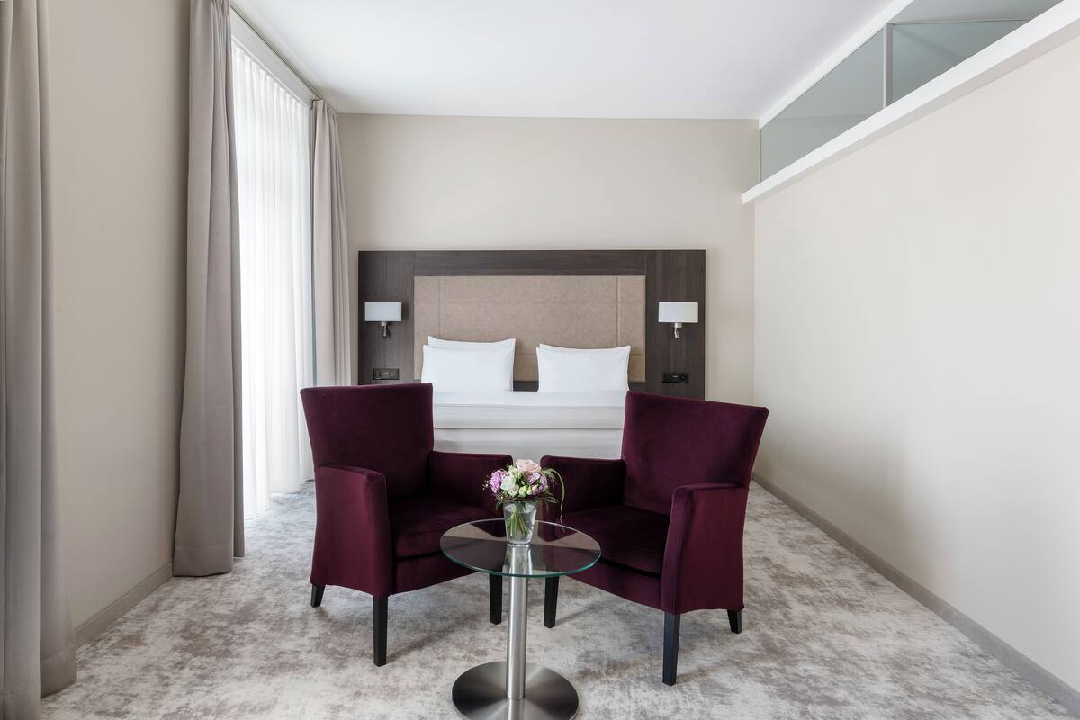 Ein Hotelzimmer mit einem Bett, zwei Sesseln, einem kleinen Tisch mit einer Vase und einer Glastrennwand.