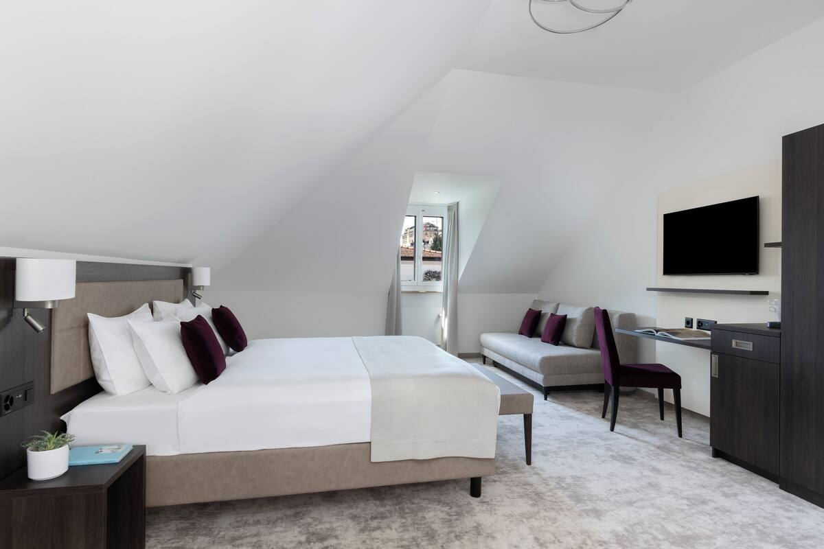 Ein modernes Hotelzimmer mit einem großen Bett, einem Schreibtisch, einem Sessel, einem Fernseher und Blick auf die Stadt.