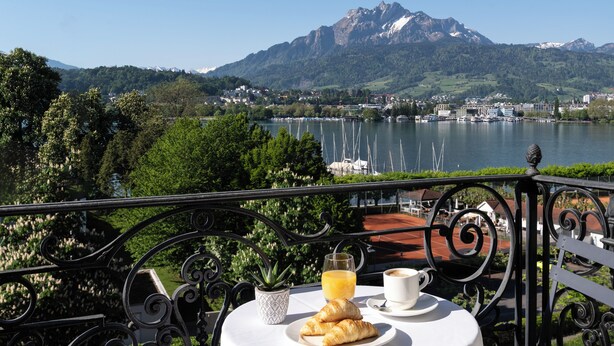 A table with croissants, a glass of orange juice, and a cup of coffee, overlooking a lake with boats and mountains in the background. - Grand Hotel Europe (Lucerne)