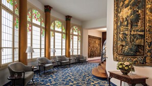 A lobby with a blue patterned carpet, four upholstered chairs, a wooden table with a vase of flowers, and large stained glass windows with floral designs. - Grand Hotel Europe (Lucerne)