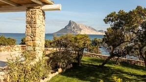 Um pilar de pedra, um telhado de madeira, uma área ajardinada e vista para o mar com uma montanha ao fundo.
