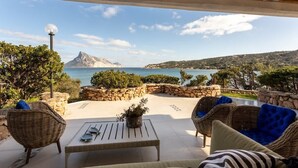 Um pátio com mobiliário de vime, uma mesa com uma planta e vista para o mar e as colinas.