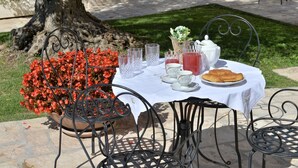 Mesa de jantar ao ar livre posta com toalha branca, copos de vidro transparente, bule, xícaras, pires, prato com croissants e um vaso com flores vermelhas.