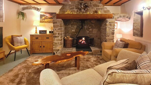 A cozy living room with a stone fireplace, wooden beams, a yellow armchair, a beige sofa, and a wooden coffee table. - 2 bedroom accommodation in Pontyberem, Carmarthenshire (Pontyberem, Carmarthenshire)
