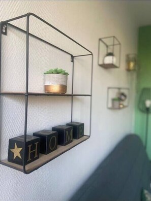 Una repisa metálica con una planta en maceta y bloques de letras que deletrean "HOME" sobre una pared con textura.