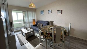 A modern bedroom with a sofa, a dining table, and chairs. There is a large window with curtains, a ceiling light, and framed pictures on the wall. - 1-Bedroom Apartment near Beach, Saint-Hilaire-de-Riez (Saint-Hilaire-de-Riez)