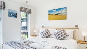 A neatly made bed with striped bedding, a window with curtains, a painting of a beach, and a bedside table with a lamp. - Carisbrooke Premier Lodge (Wootton, Nr Cowes)