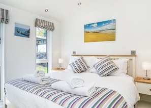 A neatly made bed with striped bedding, a window with curtains, a painting of a beach, and a bedside table with a lamp. - Carisbrooke Premier Lodge (Wootton, Nr Cowes)
