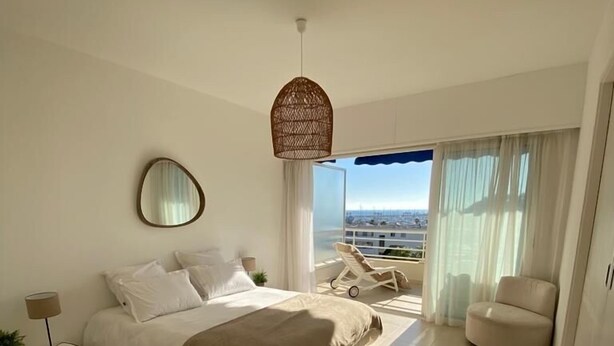 Un dormitorio moderno con una cama grande, una mesita de noche con lámpara, una lámpara colgante, una silla y vistas de la ciudad a través de amplios ventanales.