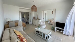 Una sala de estar moderna con sofá, mesa de centro y televisión. Se ve un comedor con mesa y sillas, y una cocina al fondo.