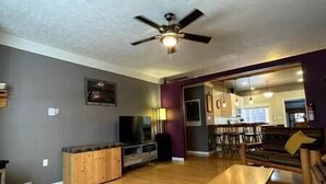 A living room with a ceiling fan, a TV, wooden cabinets, and a dining area with chairs. - Cozy, Warm & Spacious Fernie Bungalow (Fernie)