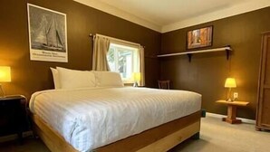 A bedroom with a large bed, a nightstand, a bedside table, a window with curtains, and a painting on the wall. - Cozy, Warm & Spacious Fernie Bungalow (Fernie)