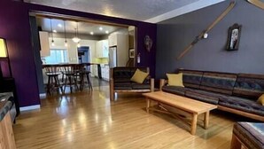 A living room with a wooden floor, a sofa, a coffee table, and a dining area with chairs and a table. - Cozy, Warm & Spacious Fernie Bungalow (Fernie)