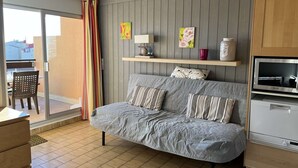 A cozy room with a grey sofa, a small table, a TV, and a window with curtains. - Quiet Studio Cabin, Close to Beach, Washing Machine, Pets OK (Cap d'Agde)