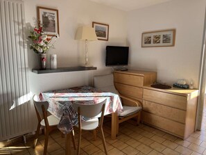 Um quarto pequeno e bem iluminado com uma mesa de jantar, cadeiras, uma TV e um armário de madeira.