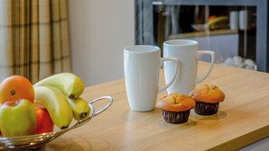 Una escena de desayuno con un bol de fruta, dos tazas blancas y dos muffins sobre una mesa de madera.
