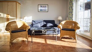Un salon comprenant un canapé, deux fauteuils en osier, une table basse en verre, un meuble, une lampe et une fenêtre avec des rideaux.
