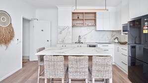 Eine moderne Küche mit weißen Schränken, einer Marmorarbeitsplatte und einem eingebauten Backofen. An der Kücheninsel stehen Korbstühle und es steht ein schwarzer Kühlschrank darin.