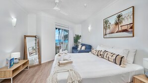 A bedroom with a large bed, a ceiling fan, a window with a view of palm trees, and a framed picture on the wall.