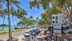 A beachfront parking area with several cars, palm trees, and a building labeled 'Alassio Palm Cove' with amenities like a coffee haven and a village gallery.
