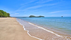A beach with a wooden pier extending into calm waters, surrounded by clear skies and a distant island.