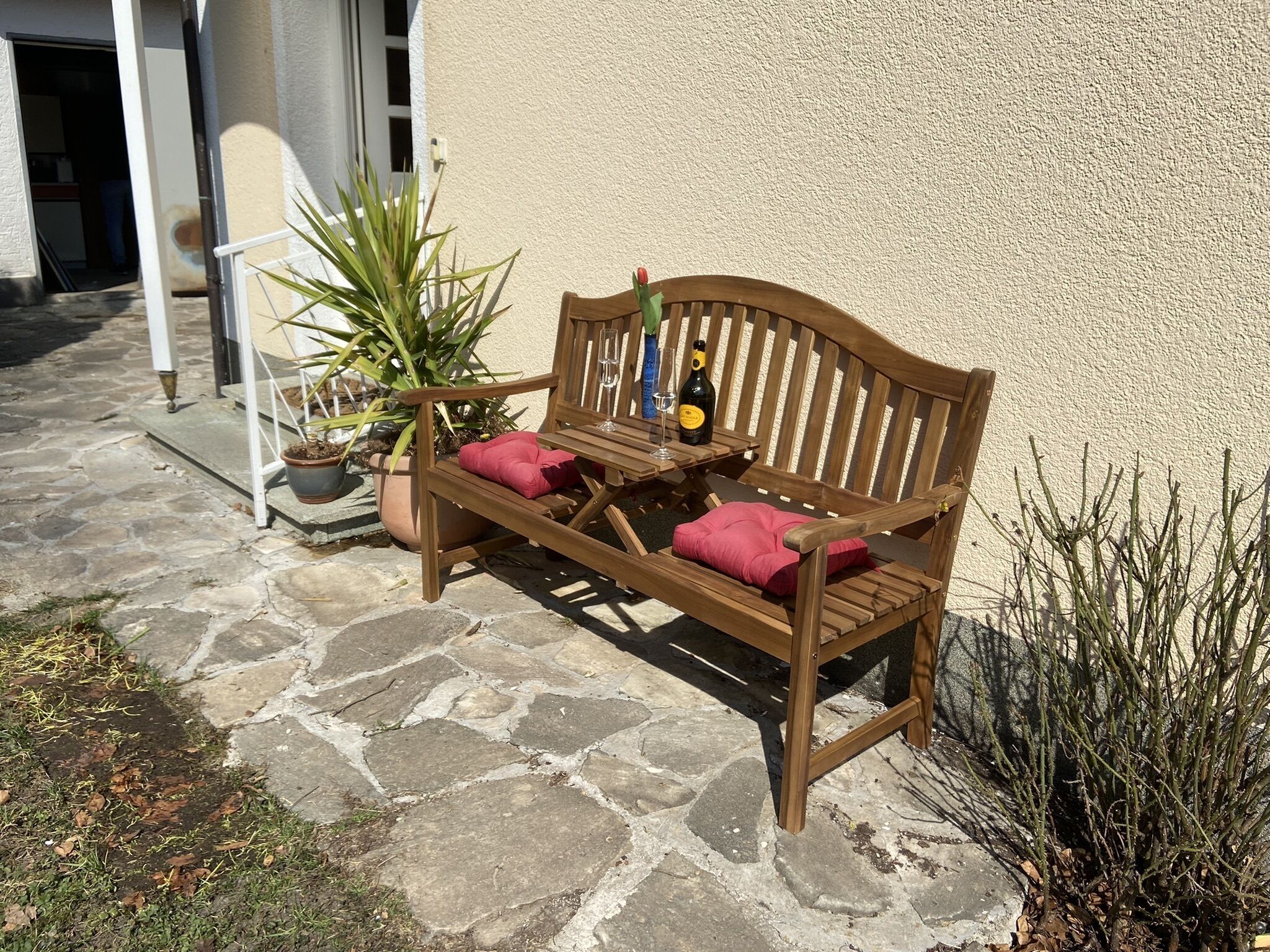 A wooden bench with cushions, a bottle of wine, and a glass on a stone patio.