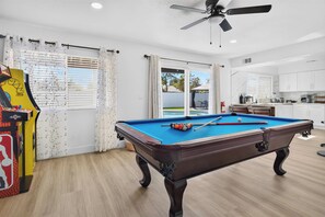 Une table de billard dans une pièce dotée d’un ventilateur de plafond, d’un coin cuisine et d’une vue sur l’extérieur, visible à travers une porte vitrée.