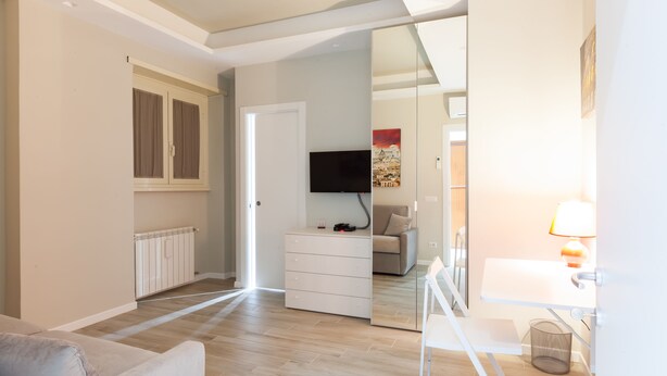 A modern, well-lit room with a wooden floor, a white dresser, a flat-screen TV mounted on the wall, and a small table with a lamp. - A Pass by St. Peter's 121 (Roma)