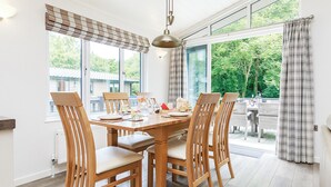A dining area with a wooden table and chairs, large windows with curtains, and a view of greenery outside. - Hartfield Watersedge Lodge (Newton Saint Loe, Nr Bath)