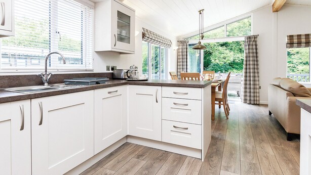 A modern kitchen with white cabinets, a granite countertop, and a dining area with a wooden table and chairs. - Hartfield Watersedge Lodge (Newton Saint Loe, Nr Bath)