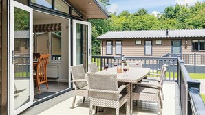 An outdoor patio with wicker furniture and a table set for dining, a sliding glass door leading to an indoor area, and a view of a building in the background. - Hartfield Watersedge Lodge (Newton Saint Loe, Nr Bath)