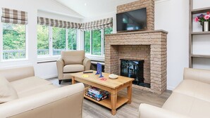 A living room with a brick fireplace, a television mounted above it, and a large window with a view of greenery. - Hartfield Watersedge Lodge (Newton Saint Loe, Nr Bath)