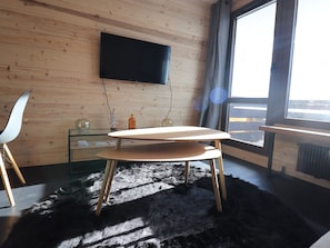 A modern room with a wooden wall, a flat-screen TV mounted above a glass table, and a large window with a view.