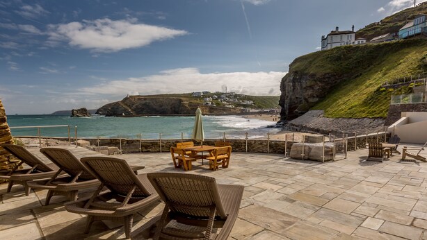 A patio with wicker chairs and tables overlooking a beach with a cliff and houses in the background. - Luxury Beachfront cottage only 20 steps from the sand with private parking/patio (Portreath)