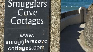 A sign for Smuggler's Cove Cottages with a coastal view. - Luxury Beachfront cottage only 20 steps from the sand with private parking/patio (Portreath)