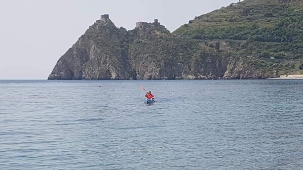 A kayaker on calm water with a rocky coastline and a castle-like structure in the background. - La Grotta (Sant'Alessio Siculo)