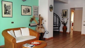 Un salon avec un canapé en osier, une table basse et un tapis à motifs. Les murs sont ornés de tableaux encadrés, et un escalier mène à une autre pièce.
