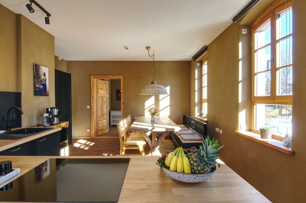 A modern kitchen with a dining area, large windows, and a fruit bowl on the table.
