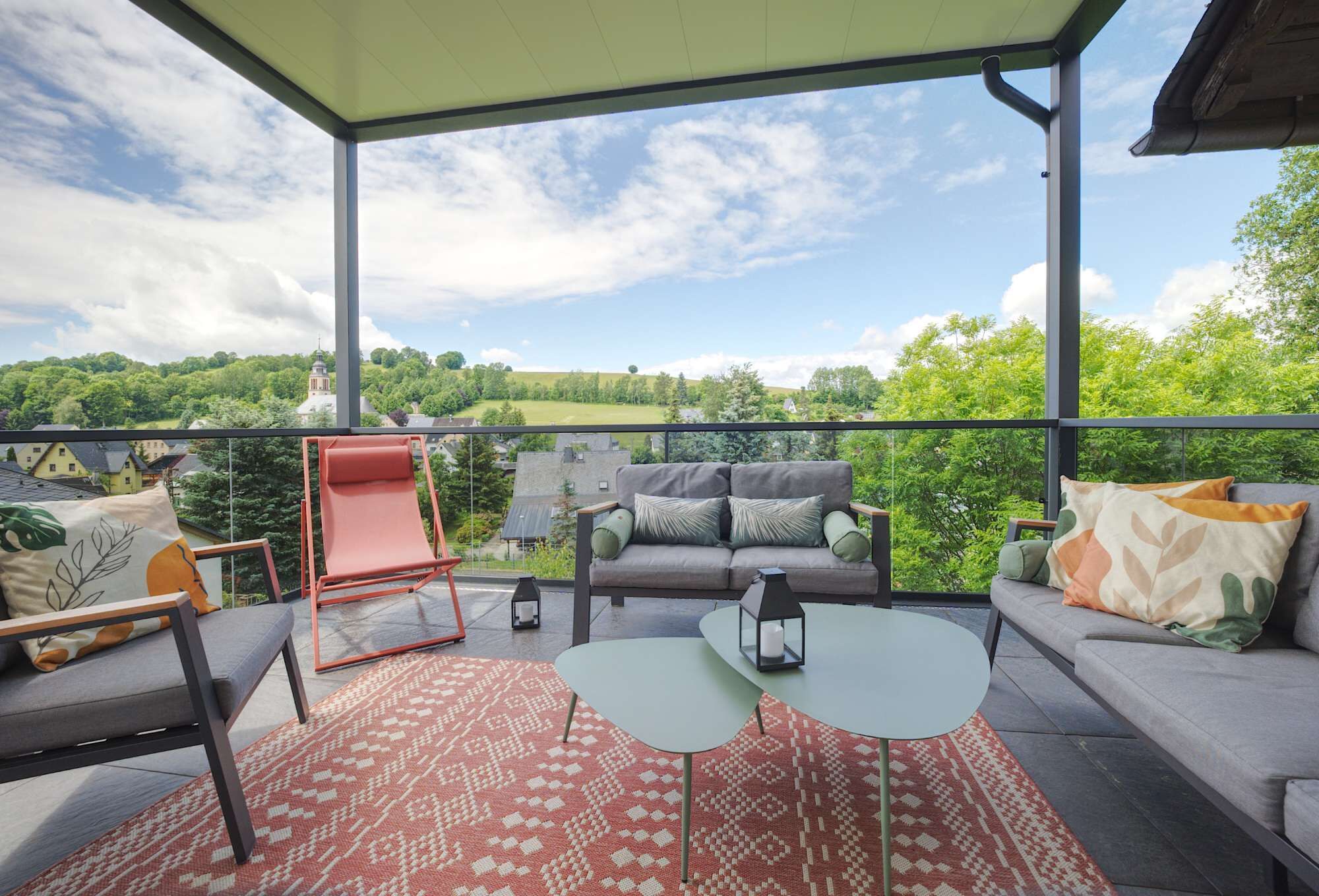 A balcony with a sofa, chairs, and a table, overlooking a green landscape and a distant building.