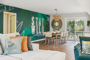 A modern living room with a green wall mural, a dining area with a wooden table and chairs, and a view of the outdoors through a large window.