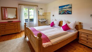 A wooden bed with white bedding and pink pillows, a wooden dresser, a wooden nightstand, a bathroom with a sink and mirror, and a painting of boats on the wall. - Spacious, renovated farmhouse in its own garden (Seehausen am Staffelsee)