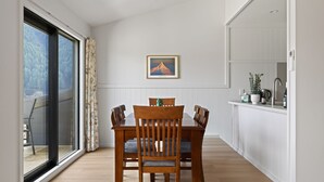 Un coin repas avec une table et des chaises en bois, une vue à travers une porte vitrée, un tableau encadré au mur, et un espace cuisine avec un évier et des placards.