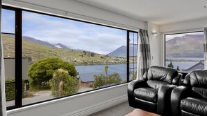 A room with a large window offering a view of a lake, mountains, and a small town. There is a black leather sofa and a wooden table. - Wonderful On Woodlands - Family friendly home (Queenstown)