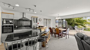 A modern kitchen with a central island, bar stools, a dining area with a table and chairs, and a view of the outdoors. - Wonderful On Woodlands - Family friendly home (Queenstown)