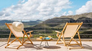 Dos sillones de madera, con un sombrero blanco para el sol en uno de ellos, una pequeña mesa redonda con una bebida y un libro, y una vista panorámica de la montaña.