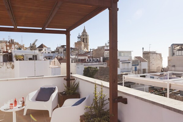 Une terrasse sur le toit agrémentée d’une pergola en bois, de deux fauteuils blancs, d’une petite table avec des boissons, et offrant une vue sur le paysage urbain, notamment le clocher d’une église.