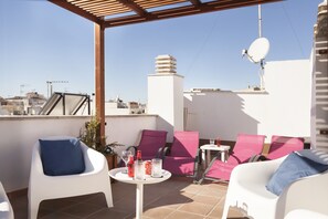 Eine Dachterrasse mit weißen und rosafarbenen Liegestühlen, einem kleinen Tisch mit Getränken und einer Satellitenschüssel.