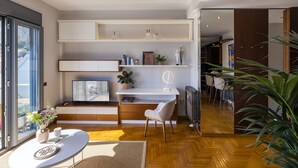 A modern living room with a white sofa, a dining table, and a built-in desk with a chair. There are shelves above the desk holding books and decorative items. A large window provides natural light, and a plant is visible in the corner. - Phos, an eclectic suite with stunning Acropolis view.<br> (Athina)
